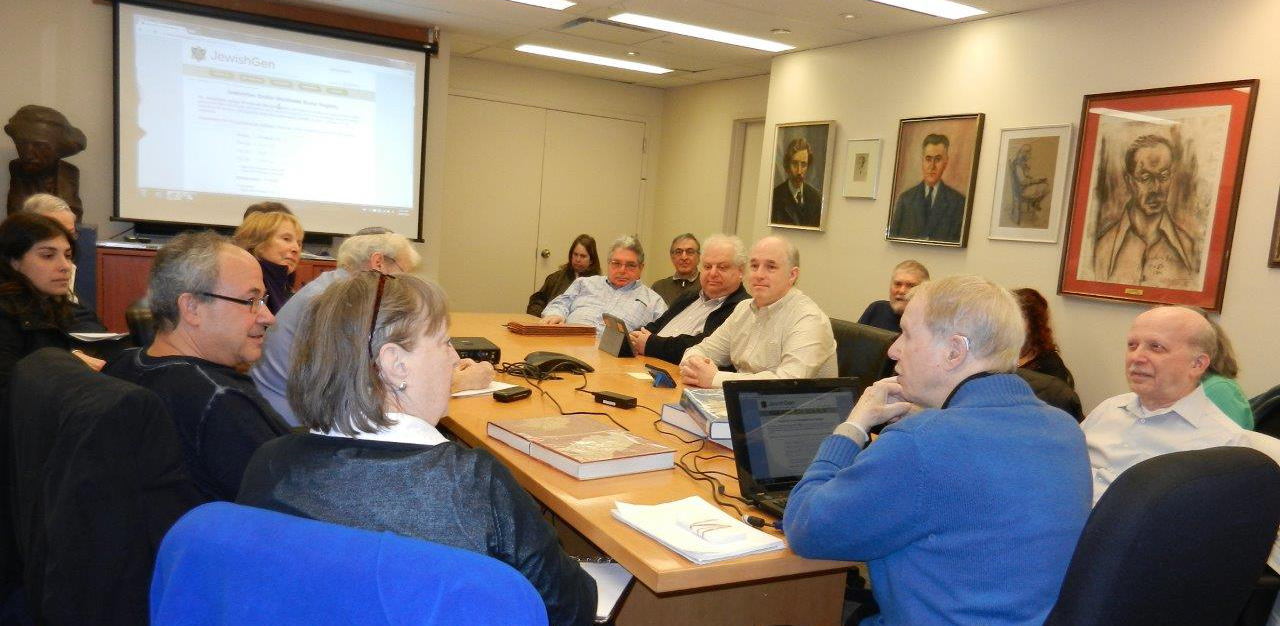 Group of people gathered around table. A laptop screen is projected on the wall.